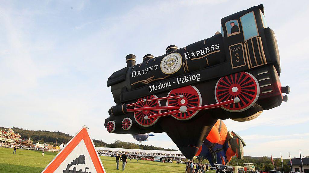 A black train with red wheels hot air balloon that is tethered near a green field.