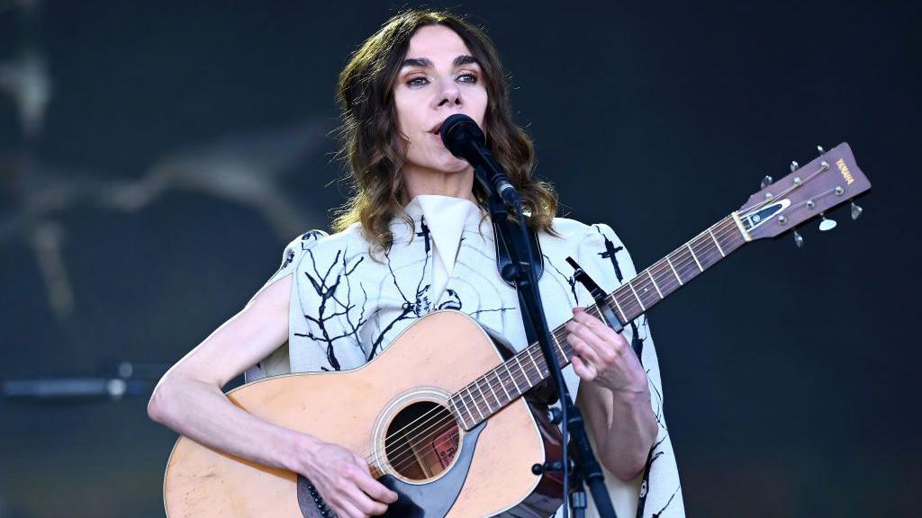 A woman with brown hair and wearing a black and white dress playing a guitar and singing into a microphone.