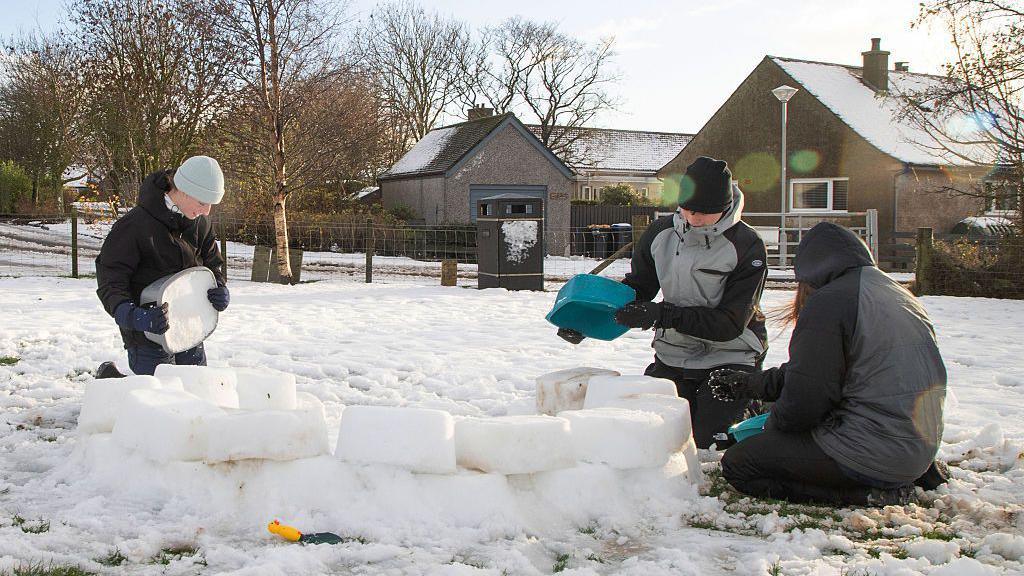 snow on the ground with three people making ice blocks and starting to make an igloo,