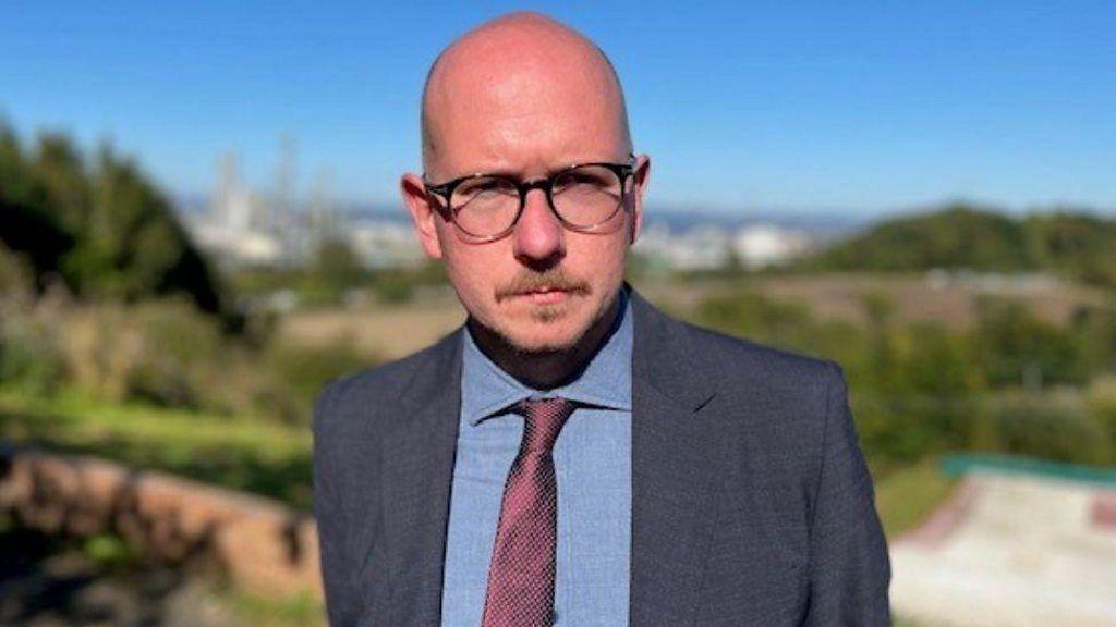 A bald man with glasses, wearing a blue suit and purple tie, looks at the camera in a medium close-up. Grangemouth refinery is in the background. 