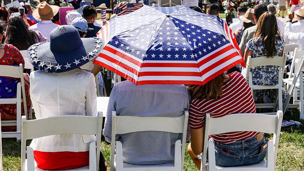 From left to right: A woman in a red dress and white jacket, her back to the camera; a man and a girl in stripey tops shelter under a US flag umbrella. All three are sitting on white wooden chairs on a grass lawn, several people sitting on matching chairs in front of them, all with their backs to the camera