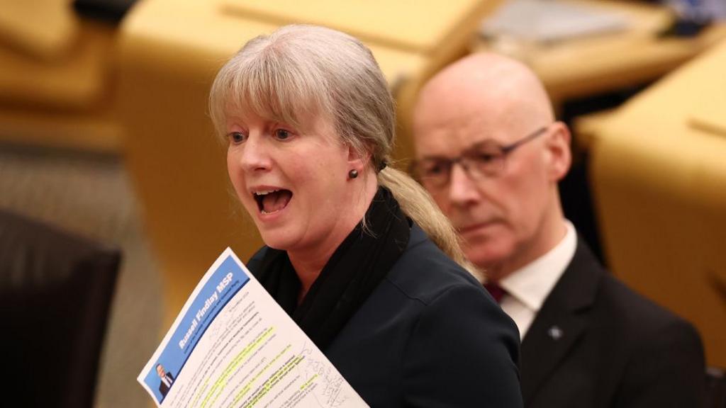 Shona Robison speaking in the Scottish Parliament with John Swinney sitting behind her. She is holding a piece of paper in her left hand.