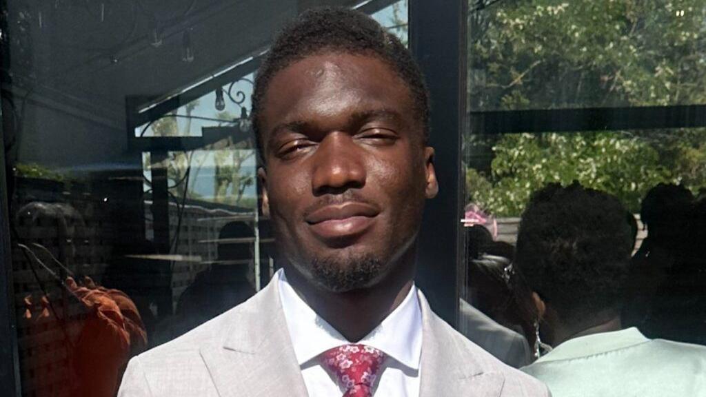 Alex Mamwa is facing the camera with his eyes closed on a sunny day, wearing a cream coloured suit jacket, white shirt and red tie