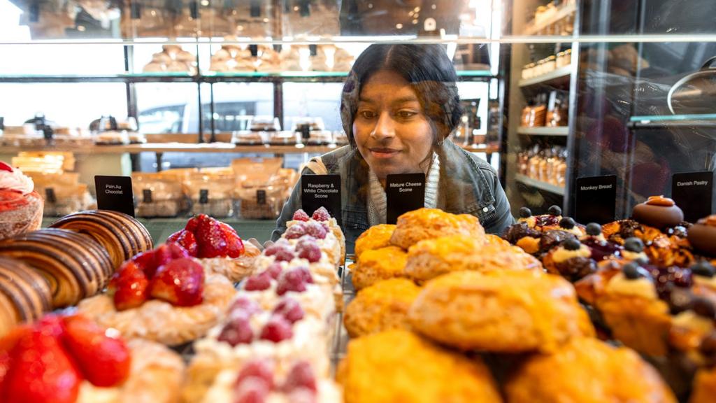 Woman peers through glass of bakery counter - a wide range of sweet items are in front of her
