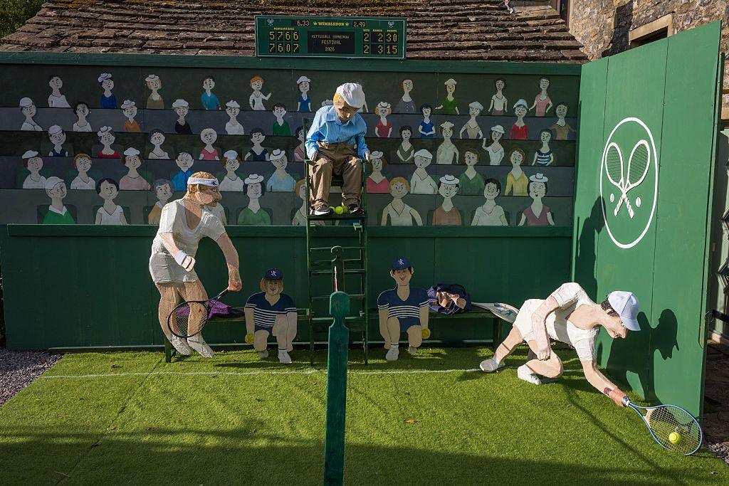 An umpire scarecrow can be seen on a tall chair, with two ball boys and two tennis players on a fake grass pitch. There is a painted piece of wood behind showing spectators painted onto it.
