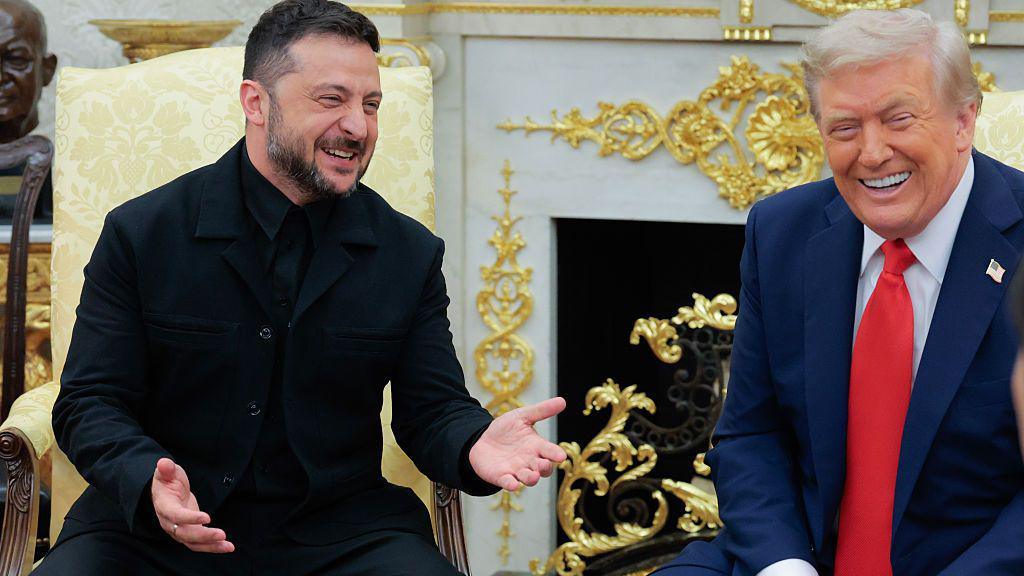 Zelensky, with short hair and a greying beard, wearing a black suit, and Trump, with grey hair, a blue jacket and a red tie. They're both sitting down and laughing, with an ornate white and gold fireplace behind them.