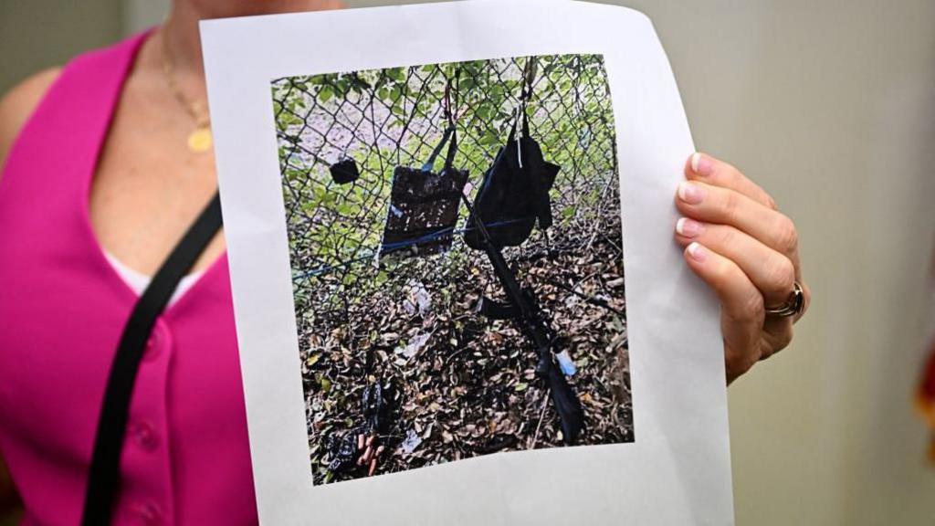 A printed photo held up by a person wearing a pink top shows the rifle and other items, that look like bags hanging on a fence, found near where a suspect was discovered during a press conference regarding an apparent assassination attempt of former President Donald Trump on September 15, 2024, in West Palm Beach