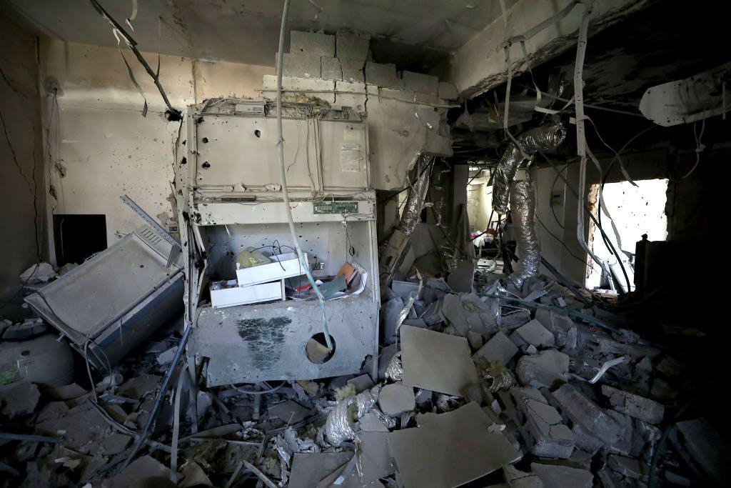 There is rubble on the floor, the walls are damaged, a large piece of equipment has been destroyed and cables hang from the ceiling.