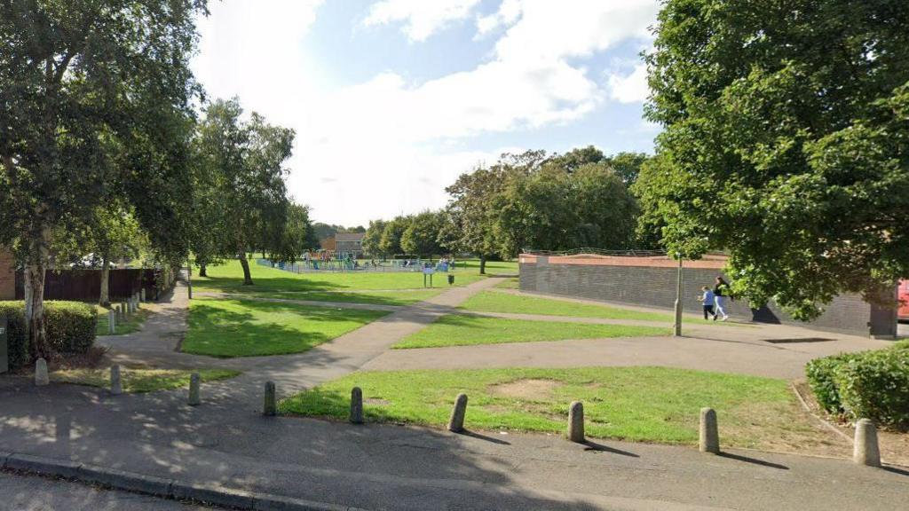 The park area by Ruscote Arcade on a sunny day. Some people are seen walking on the paths.