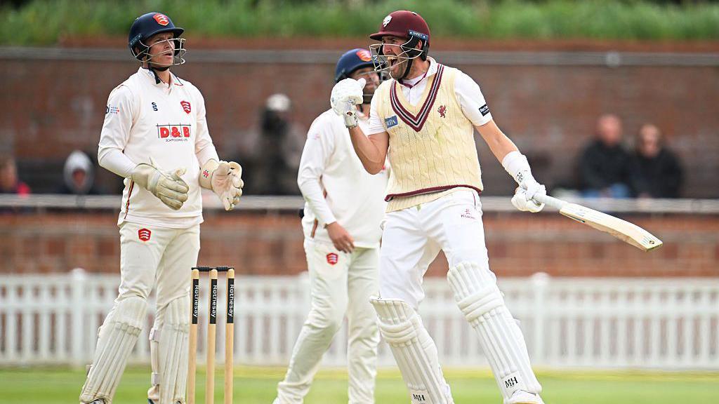 Three men in cricket gear are standing on a pitch. One of them is laughing and raising a fist.