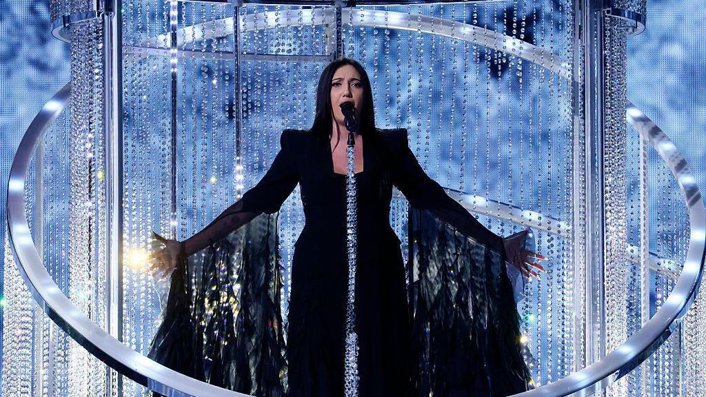 Israel's Eurovision 2025 entrant, Yuval Raphael, sings on stage surrounded by giant hoop chandeliers at the contest in Basel, Switzerland in May. She is wearing a dark dress which has long, frilly sleeves extending to the floor.