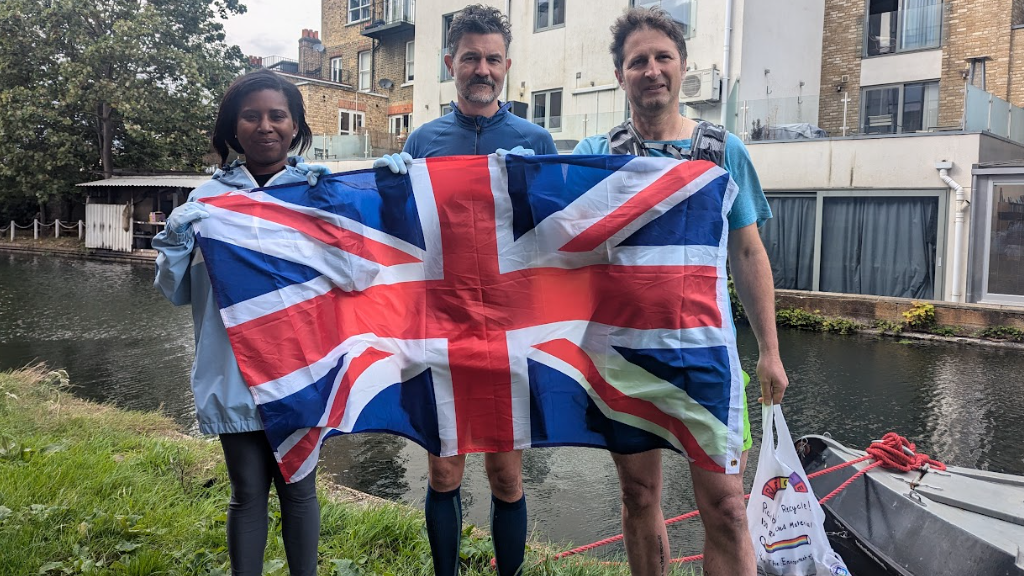 A woman and two men on a canal side in jogging gear and gloves, with one holding a rubbish bag and all standing behind a large Union Jack which is draped across them.