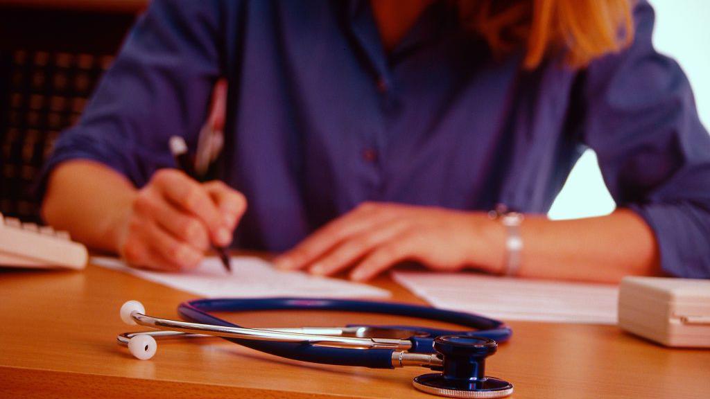A woman with blonde hair and a blue shirt is sitting at a desk and writing. In front of her on he desk is a stethoscope