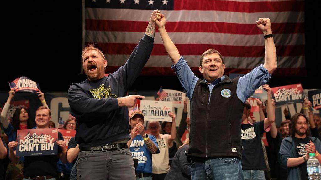 Two men in jeans and sweatshirts clasp their hands and raise them high before an American flag and small cheering crowd holding signs that read "A Senator Money Can't Buy"
