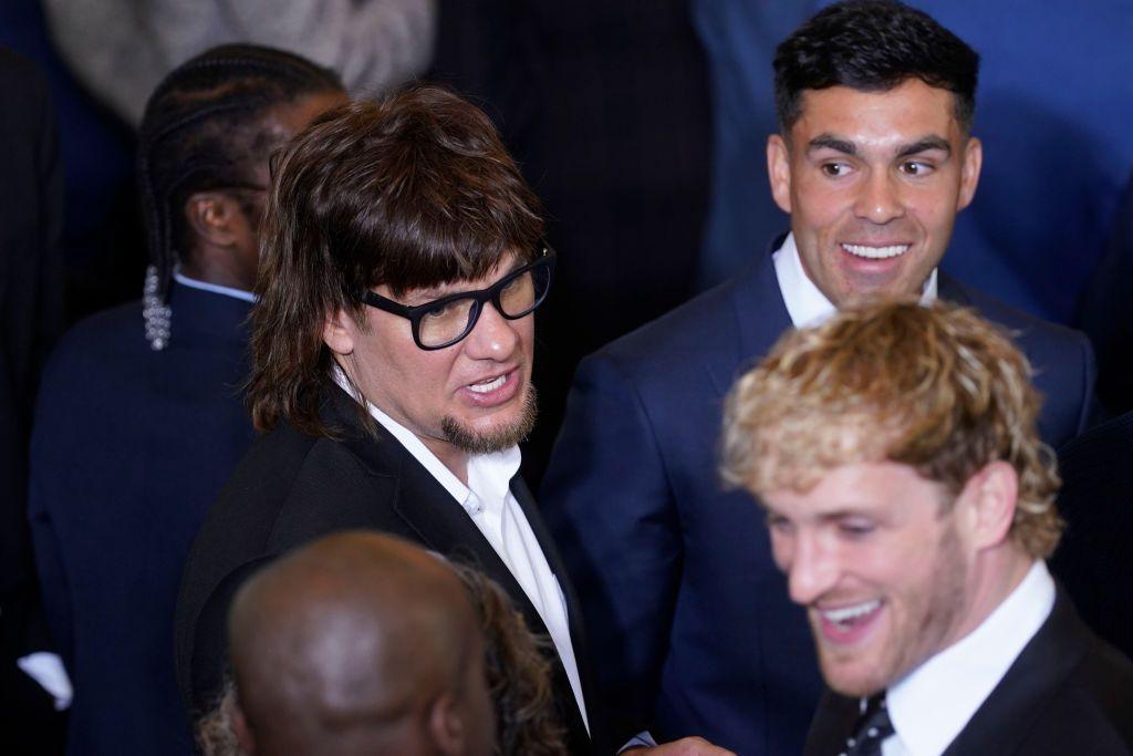 A man with a mullet and glasses stands next to a man with dark hair and a man with blond shaggy hair. They all wear suits.