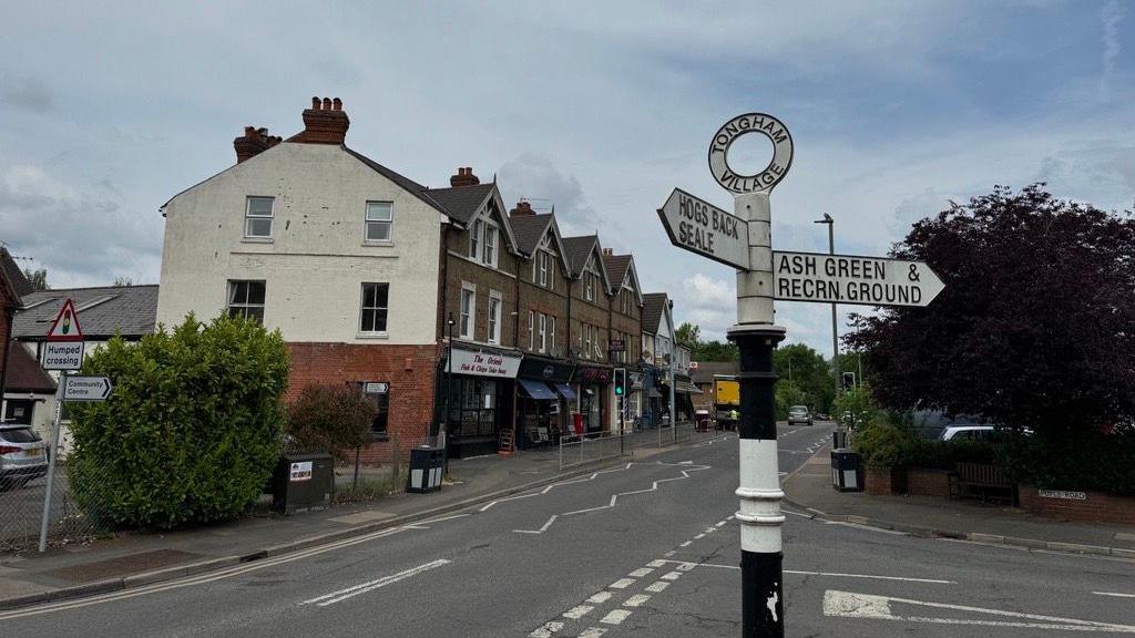 A road sign in the middle of Tongham, pointing to Hogs Back and Seale in one direction, and Ash Green in the opposite direction. The main road with a row of shops and houses is behind it, as is a T-junction.