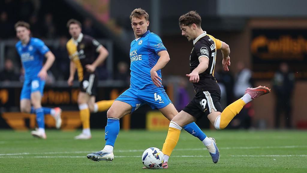 Archie Collins, wearing a blue Posh kit, prepares to tackle the ball from Dylan Williams of Burton Albion, wearing a black and yellow kit on the pitch.