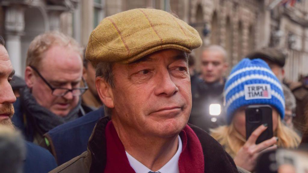 Nigel Farage in a flatcap and scarf surrounded by people 