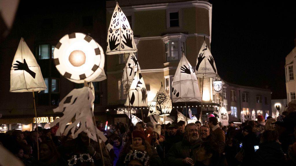 A parade of smaller lanterns held up by a crowd making its way through the streets. They are passing the buildings that line the streets in the night time scene.