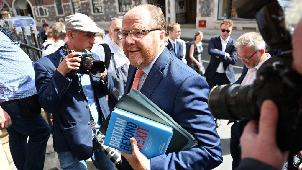 George Freeman is holding a file and a book while surrounded by photographers.