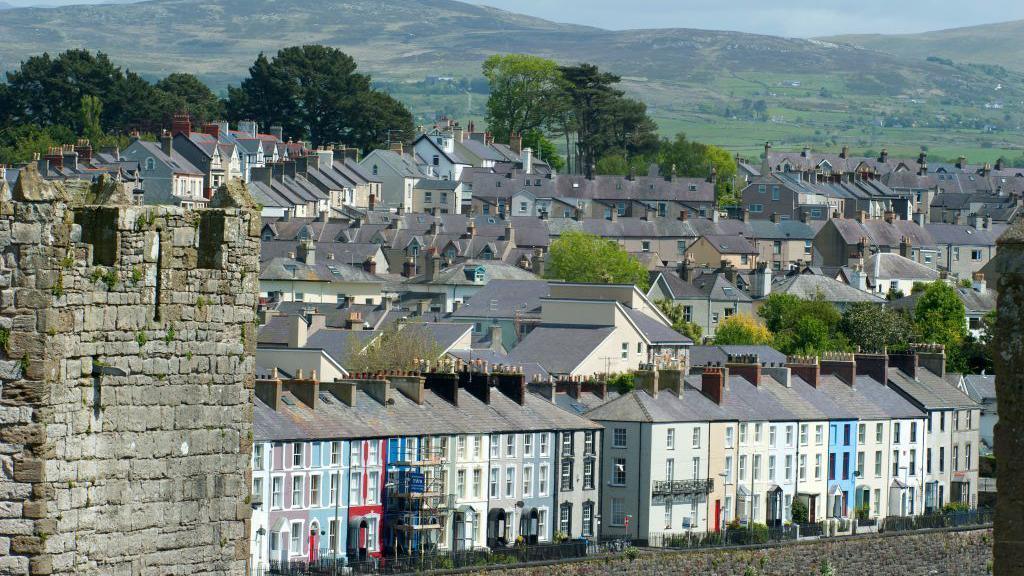 View from the medieval castle of Caernarfon on the village. Multicoloured three story terraced houses can be seen at the forefront of the image, with the corner of a medieval castle on the left of the photograph. Roofs of homes cover the mountain behind the line of houses. A large mountain can be seen behind the homes. 