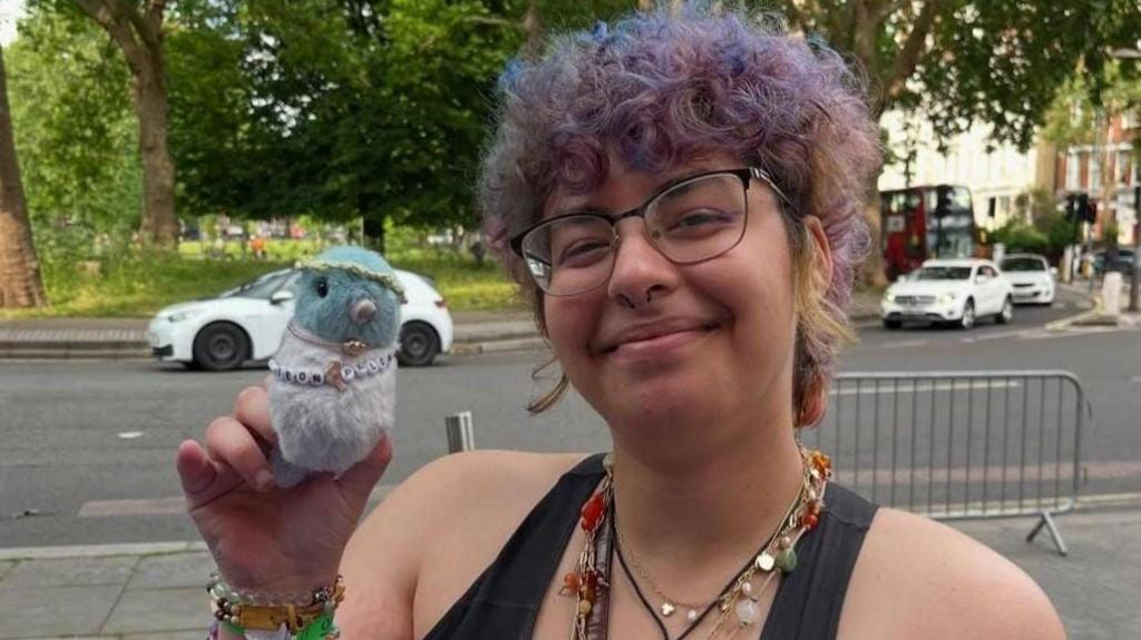 Student Alex Mustafa smiles on the street while holding a toy pigeon. She has short purple hair and is wearing lots of beaded bracelets.