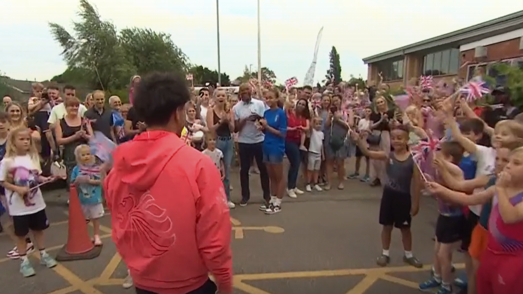 Jake Jarman in a red tracksuit top is cheered on by crowds, many of whom are waving Union Jacks, as he walks to his gymnastics club