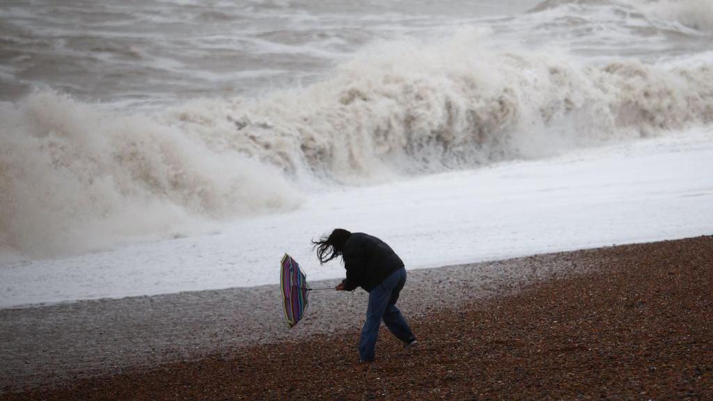 A woman holds on to her broken umbrella as she has walks along a beach in stormy weather 