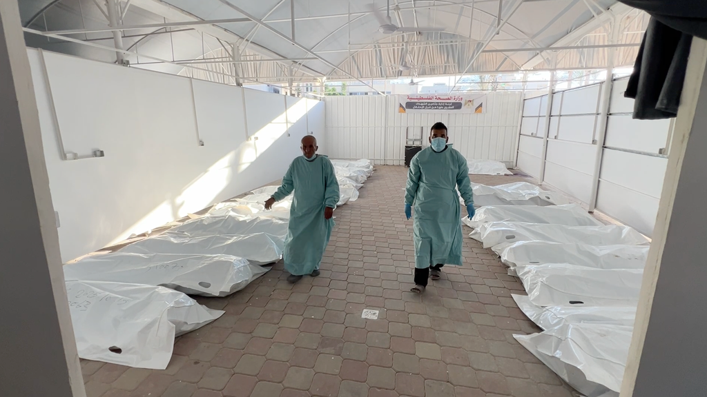 Two men in blue surgical gowns are seen inside a room with multiple white draped shapes on the floor, which contain bodies.
