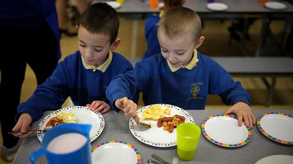 Boys wearing school uniform eat a plate of eggs and sausages in a school canteen.