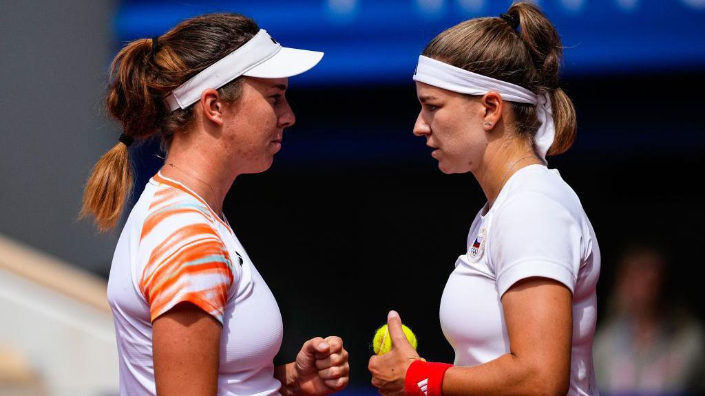 Linda Noskova and Karolina Muchova stand face to face while representing the Czech Republic at the Olympics, with Muchova holding a tennis ball in her left hand