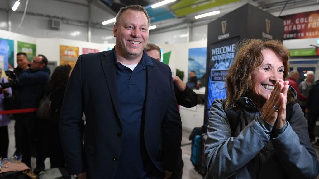 CEO Mikael Peterson. He is tall with dark hair and smiling wearing a dark blue blazer, dark blue polo shirt and white t-shirt. A woman is standing next to him with dark hair wearing a clue coat. She is excitedly smiling holding her hands together.