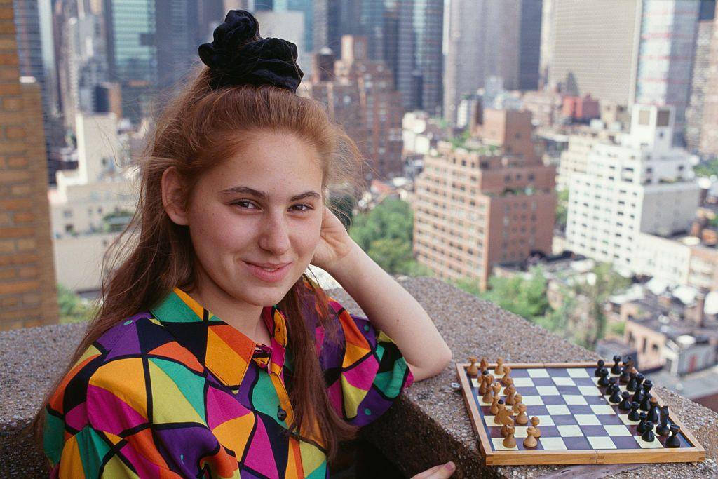 Judit Polgar as a teenager pictured on a balcony of a high rise building in a big city. She is standing next to a small chess board, wearing a colourful shirt. Her hair is in a high ponytail with a large scrunchie. She is smiling at the camera.