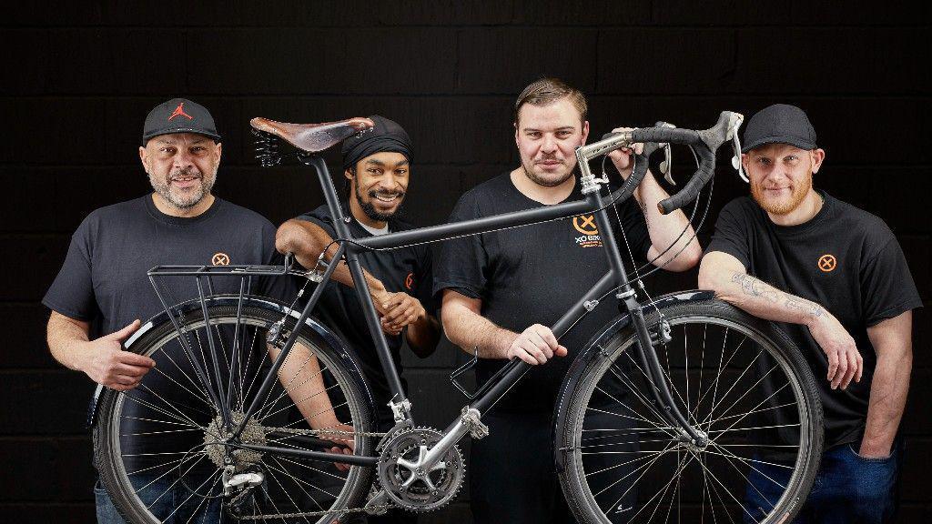 Four men, all wearing black t-shirts, stand behind a racing bike and in front of a black background.