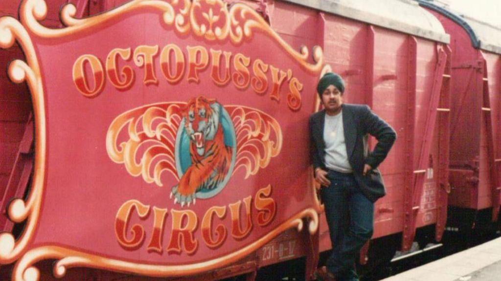 A young Del is standing on the film set next to the red train There is a sign on it which reads 'Octopussy's Circus'. Del is leaning against it with one hand on his hip. He is wearing jeans, a turquoise turban, gold necklace, grey top and blue and black striped blazer.