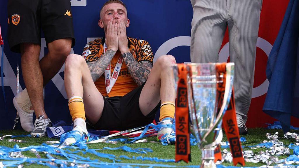 Oli McBurnie sitting on the ground in his amber Hull City shirt with his hands over his mouth in contemplation with the play-off trophy in front of him