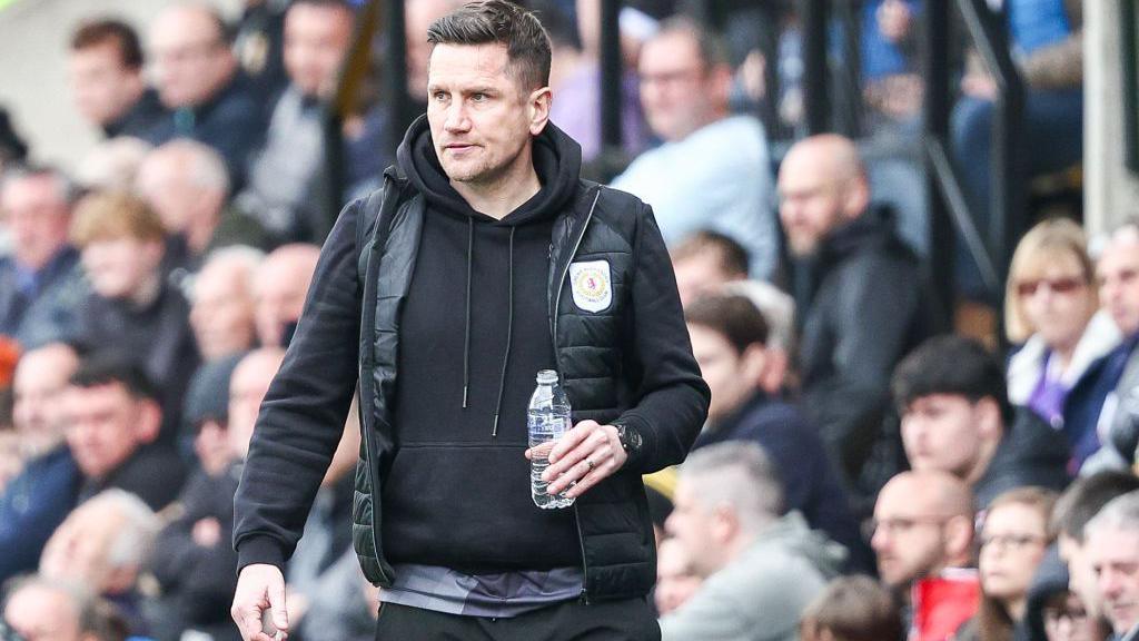 Lee Bell holds a bottle of water in his hand as he walks along the touchline watching his Crewe team in aciton