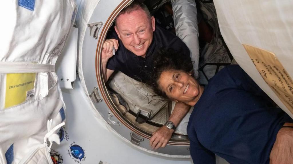 Two Nasa astronauts - Butch Wilmore and Suni Williams -  in a small vestibule area in a spacecraft, both smiling at camera