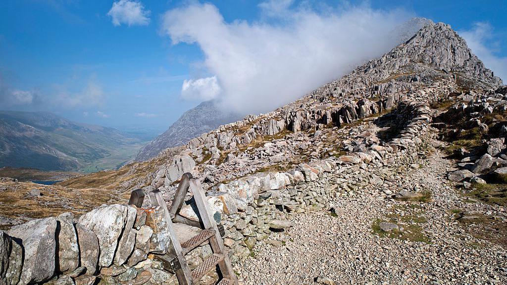 The peak of a mountain, with a rocky path, a wooden ladder and clouds hugging the summit