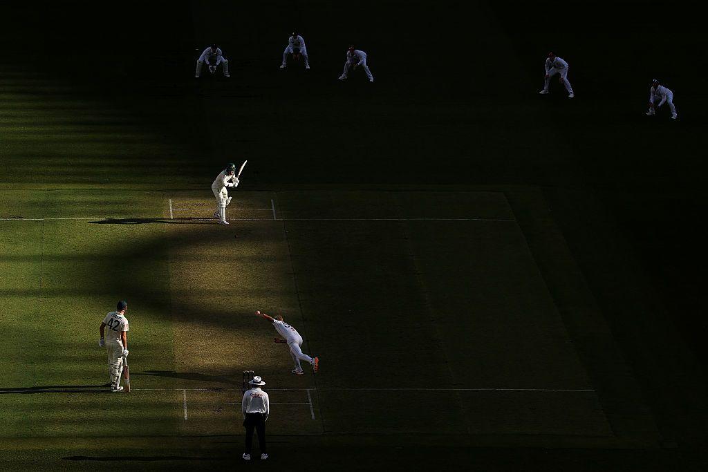 A cricket bowler delivers a ball under dramatic shadows as the batter defends, surrounded by slips and close fielders standing in bright light against a mostly dark outfield.
