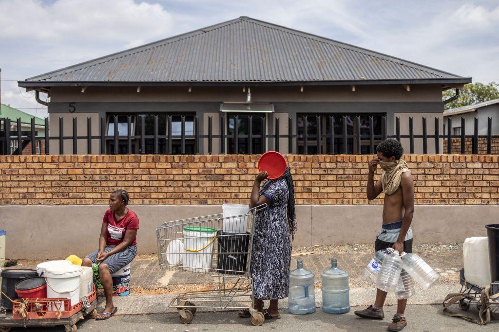 People queue for water in Johannesburg. One person is sitting at the roadside with a number of containers, another stands, holding on to a shopping trolley containing vessels and also covering her face; a third stands with a number of large containers in his hand.