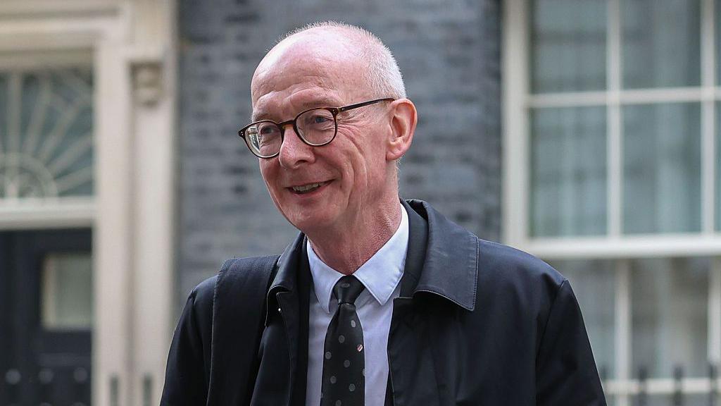 A smiling Pat McFadden stood in front of Number 10, Downing Street. He is wearing a black suit with a blue shirt and a black tie with grey polka dots with a black rain jacket over the top and a bag over his shoulder.