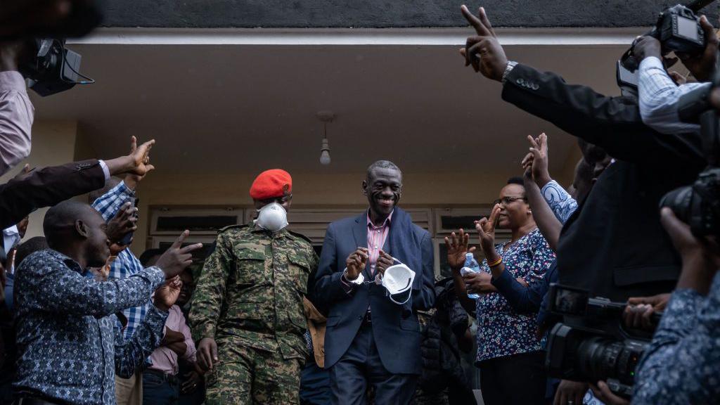 A smiling Kizza Besigye in a blue suit emerges from court accompanied by a man in fatigues and a red beret, wearing a face mask. Supporters are around him waving their fingers in a peace sign. 