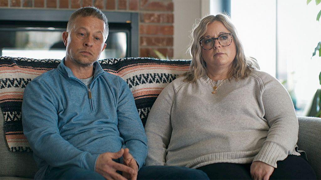 Picture of Brad and Kari Boettler. They are sitting together on a sofa in their living room, with glass patio doors and an exposed brick wall behind them. Brad has short fair hair and is wearing a pale blue jumper with a zip at the neck. Kari has tortoiseshell glasses, shoulder-length wavy blonde hair, and is wearing an oatmeal-coloured crew neck jumper with a gold necklace.
