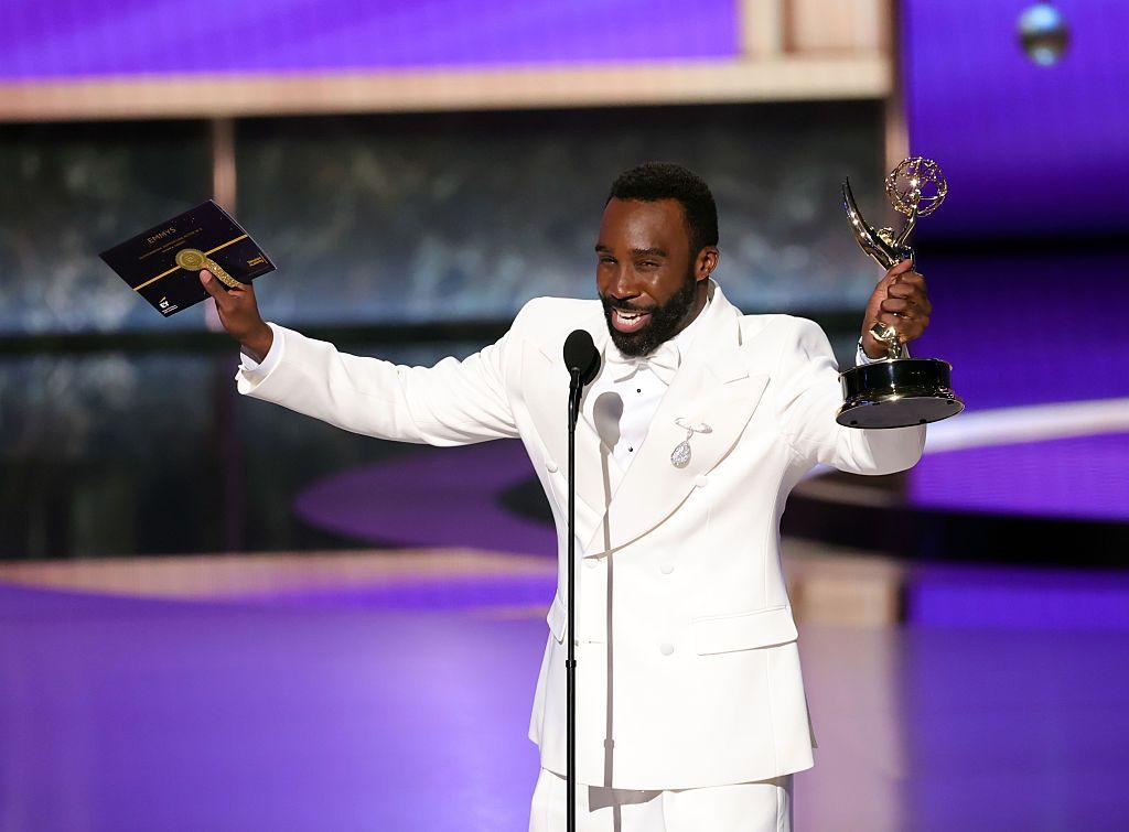Actor Trammell Tillman wears a white shirt and tuxedo and stretches his arms out while holding his Emmy Awards trophy in one hand and an envelope in another.
