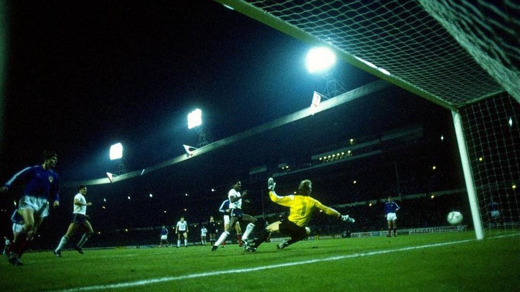 Viv Anderson scores for England against Yugoslavia at Wembley in November 1986. He is in the six-year box as the goalkeeper dives to his right in vain. The sweeping stand and floodlights of the old Wembley Stadium form the background.