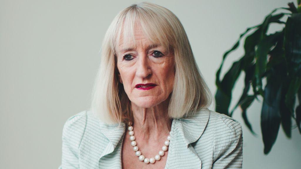 Prof Janice Rymer speaking to the camera in a white-walled room. She has grey, straight, shoulder-length hair with a fringe and is wearing a dark pink lipstick. She has a light blue suit jacket and a large pearl necklace. There is a plant positioned behind her.