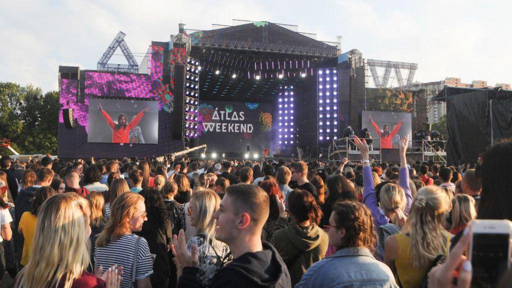 A crowd of young people stand in front of a big music stage at a music festival. On the screen behind the stage are the words "Atlas Weekend"