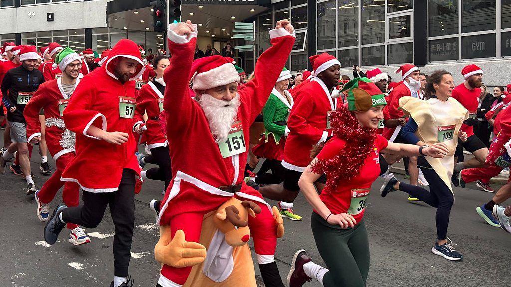 A group of people wearing red Santa suits and other festive costumes running down a hill. A man at the front has his arms aloft.