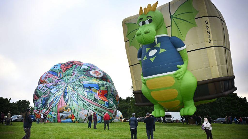 Slick the Dragon hot air balloon tethered on a field. The hot air balloon shows a green dragon with a blue t-shirt and a crate of pallets attached to its back. To the left is a colourful hot air balloon which is being inflated on the grass.